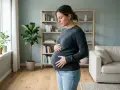 Gravid kvinna med händerna på magen, kanske känner hon av mycket sammandragningar. Hon står i ett ljust rum med en soffa och bokhylla i bakgrunden.