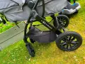 En barnvagn från Kinderkraft 3 in 1 står på gräset. Den har en svart ram, grå tygdelar och en bilbarnstol.