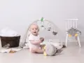 Glad baby leker under en babygym båge med mjuka fågelleksaker. En stol med en uggla och en träbalja med handdukar i bakgrunden.