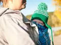 Ett barn i bärsjäl tittar fram bakom en vuxen. Barnet har en grön mössa med tofs och en randig halsduk.