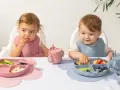 Två små barn äter från varsin tallrik ur ett barnservis i silikon. En rosa och en blå.