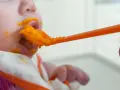 En bebis får mat på en sked, med orange puré runt munnen. En liten mage växer, och snart är det dags för mer mat.