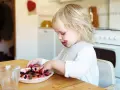 Ett barn med blåbärsfläckar i ansiktet leker med sitt mellanmål, en tallrik full med blåbär.