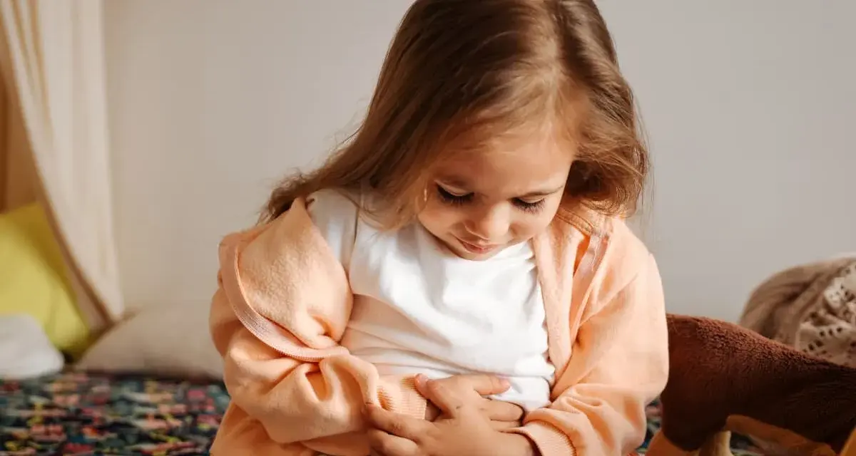 Liten flicka med magont, kanske bebis diarre. Hon sitter på en säng med händerna på magen.