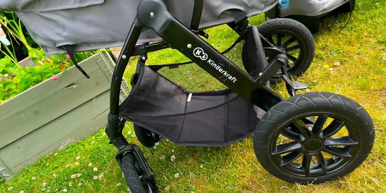 En barnvagn från Kinderkraft 3 in 1 står på gräset. Den har en svart ram, grå tygdelar och en bilbarnstol.
