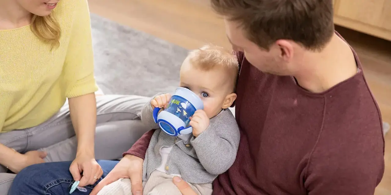 En bebis dricker ur en blå nuk magic cup medan föräldrarna tittar på.