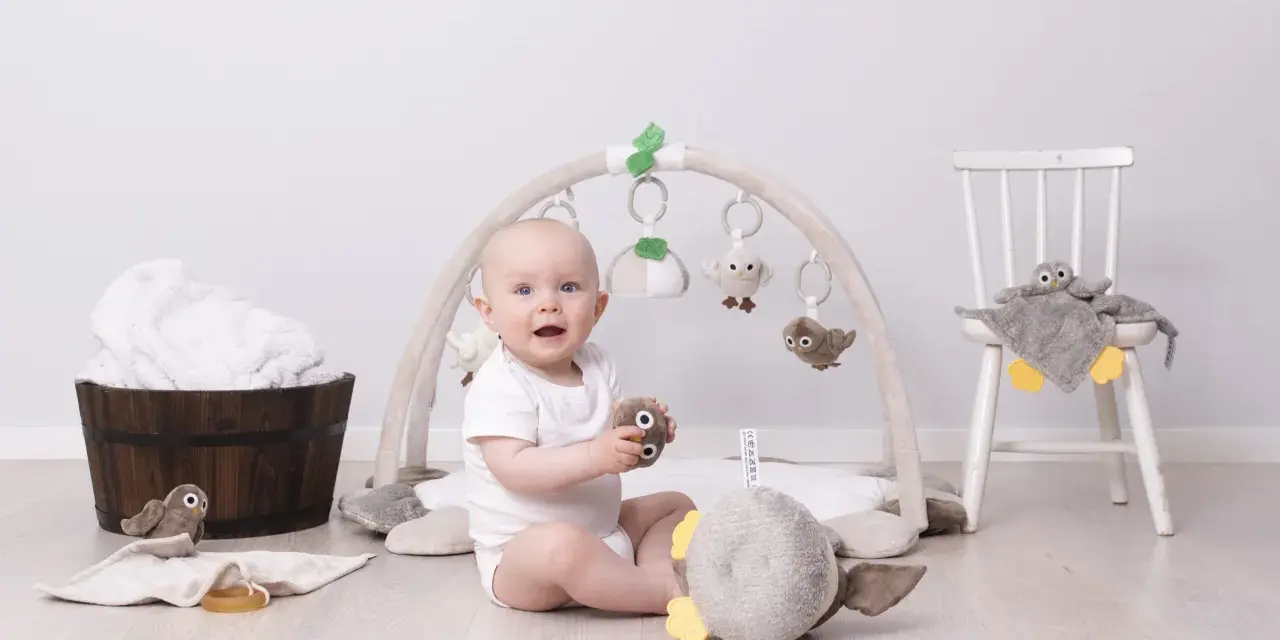 Glad baby leker under en babygym båge med mjuka fågelleksaker. En stol med en uggla och en träbalja med handdukar i bakgrunden.