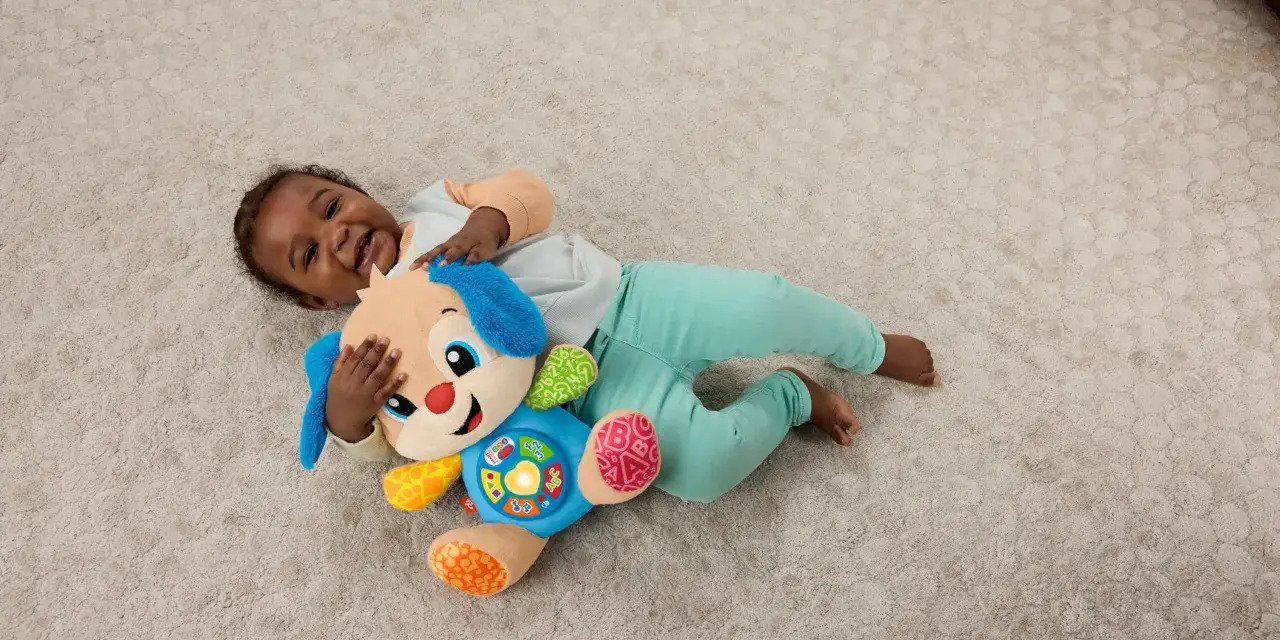 Ett litet barn skrattar och kramar sin Fisher Price hund på en ljus matta. Hunden har blå öron och en färgglad mage med knappar.