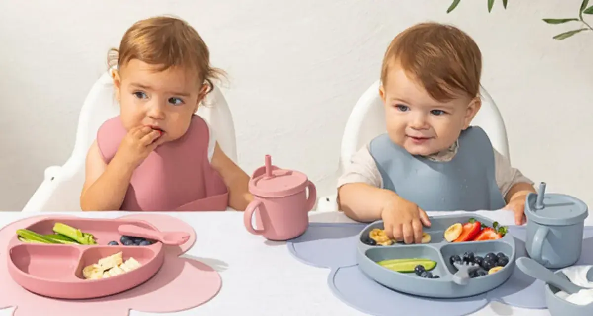 Två små barn äter från varsin tallrik ur ett barnservis i silikon. En rosa och en blå.
