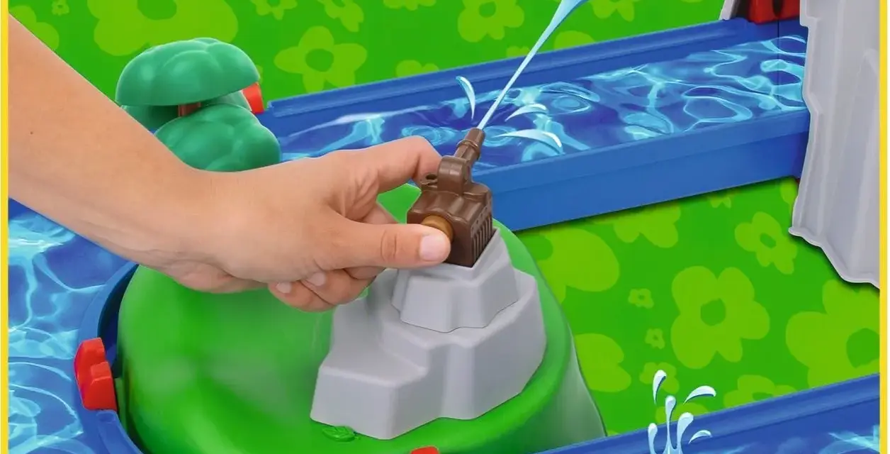 En hand aktiverar en vattenpump på en Aquaplay mountain lake-lekplats, som sprutar vatten genom en blå kanal.