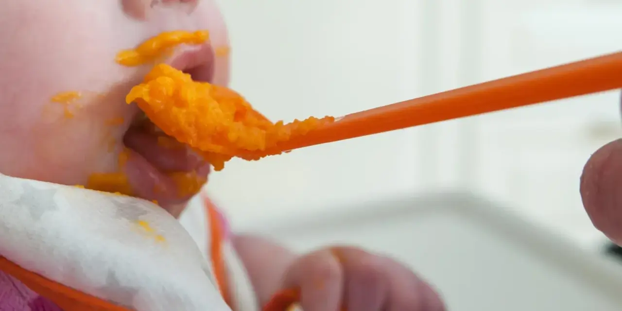 En bebis får mat på en sked, med orange puré runt munnen. En liten mage växer, och snart är det dags för mer mat.