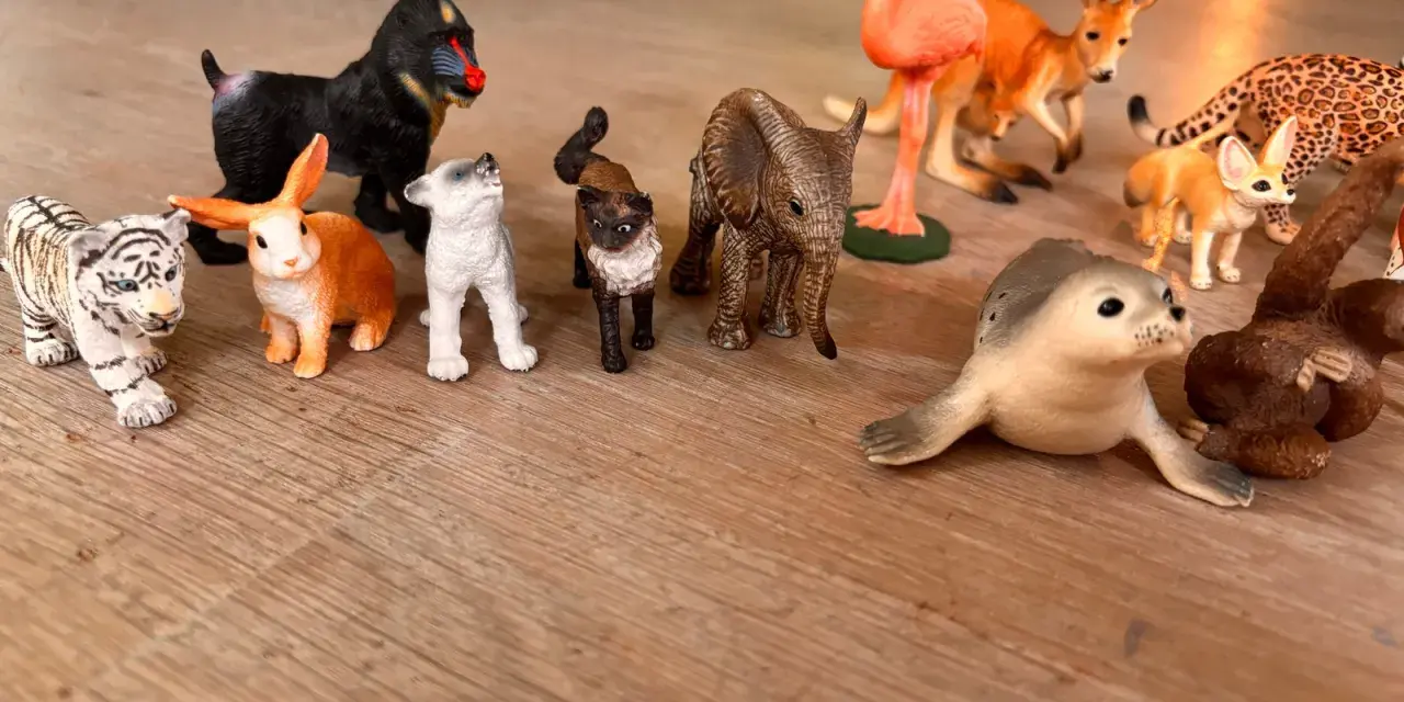 En samling schleich vilda djur leksaker på ett trägolv: tiger, apa, kanin, hundvalp, katt, elefant, flamingo, känguru, fennek, säl och en apa.