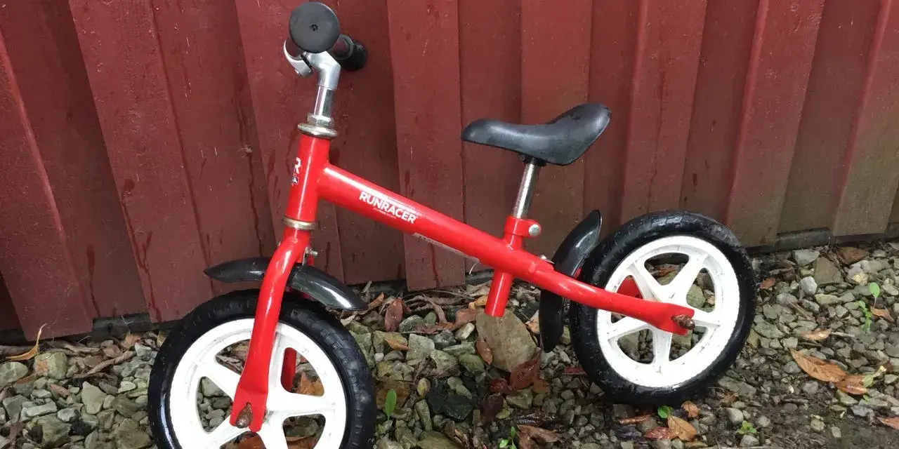 En röd stiga springcykel står lutad mot en röd trävägg. Den har vita fälgar och svarta däck.