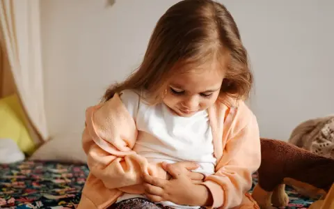 Liten flicka med magont, kanske bebis diarre. Hon sitter på en säng med händerna på magen.
