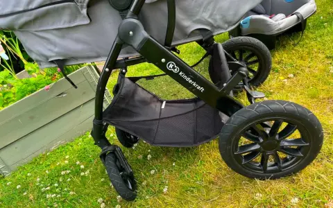 En barnvagn från Kinderkraft 3 in 1 står på gräset. Den har en svart ram, grå tygdelar och en bilbarnstol.