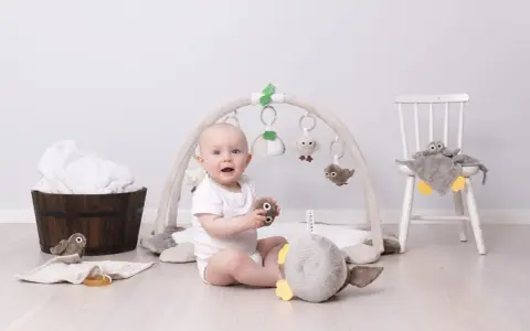 Glad baby leker under en babygym båge med mjuka fågelleksaker. En stol med en uggla och en träbalja med handdukar i bakgrunden.