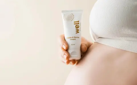 En hand håller en tub med "well Nipple & Barrier Cream", liknande Medela Purelan, nära en gravid mage.