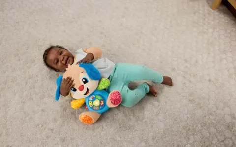 Ett litet barn skrattar och kramar sin Fisher Price hund på en ljus matta. Hunden har blå öron och en färgglad mage med knappar.