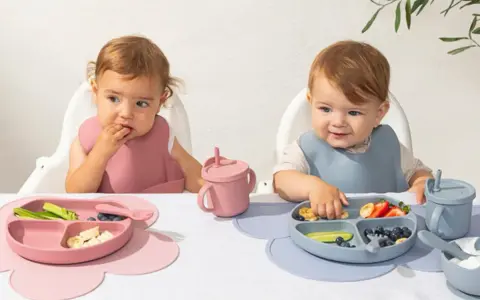 Två små barn äter från varsin tallrik ur ett barnservis i silikon. En rosa och en blå.