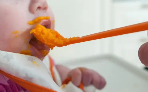 En bebis får mat på en sked, med orange puré runt munnen. En liten mage växer, och snart är det dags för mer mat.
