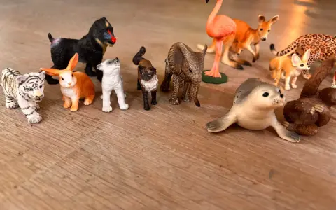 En samling schleich vilda djur leksaker på ett trägolv: tiger, apa, kanin, hundvalp, katt, elefant, flamingo, känguru, fennek, säl och en apa.