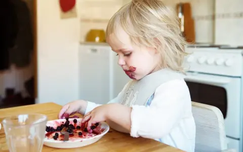 Ett barn med blåbärsfläckar i ansiktet leker med sitt mellanmål, en tallrik full med blåbär.