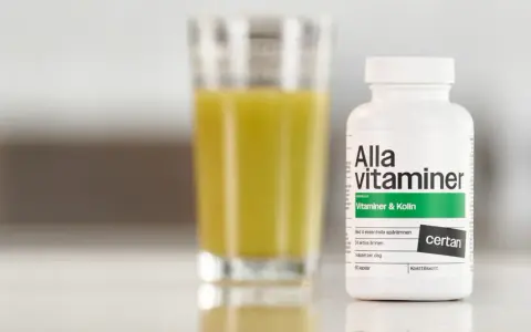 En flaska "Alla vitaminer" med texten "Vitaminer & Kolin" står bredvid ett glas med gul vätska. Perfekt för den som behöver extra vitaminer under graviditeten.