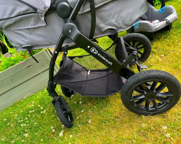 En barnvagn från Kinderkraft 3 in 1 står på gräset. Den har en svart ram, grå tygdelar och en bilbarnstol.