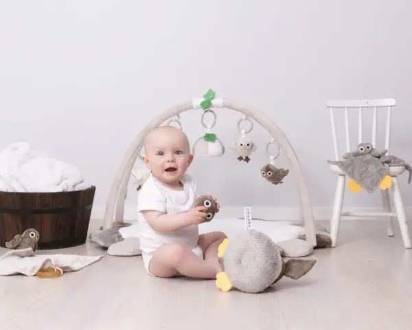 Glad baby leker under en babygym båge med mjuka fågelleksaker. En stol med en uggla och en träbalja med handdukar i bakgrunden.