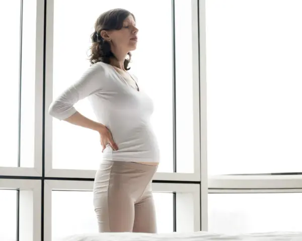 Gravid kvinna med ont i mitten av ryggen, håller sig mot ländryggen. Hon står framför ett stort fönster.