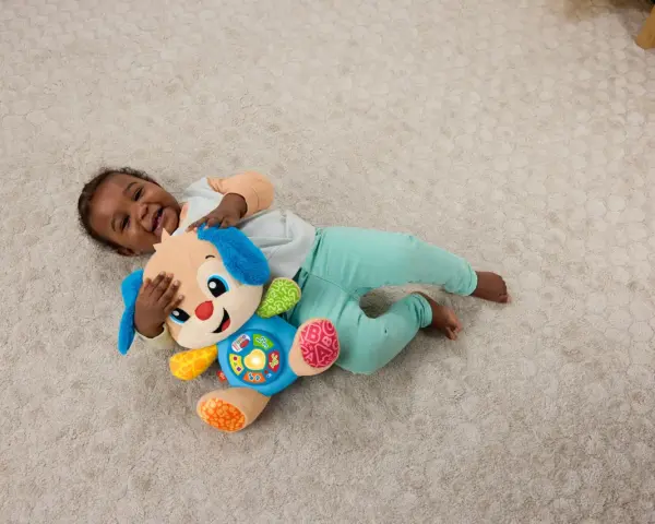 Ett litet barn skrattar och kramar sin Fisher Price hund på en ljus matta. Hunden har blå öron och en färgglad mage med knappar.