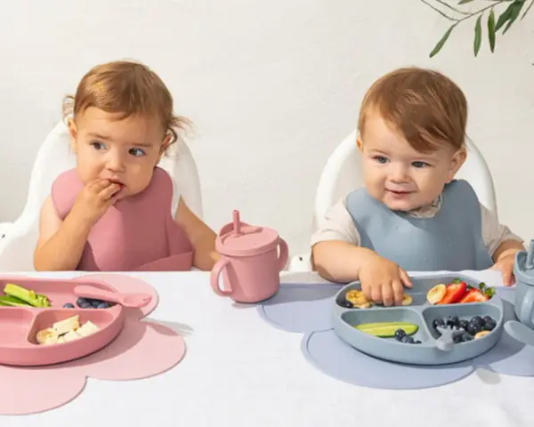 Två små barn äter från varsin tallrik ur ett barnservis i silikon. En rosa och en blå.
