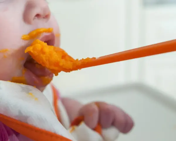 En bebis får mat på en sked, med orange puré runt munnen. En liten mage växer, och snart är det dags för mer mat.