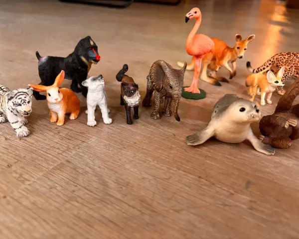 En samling schleich vilda djur leksaker på ett trägolv: tiger, apa, kanin, hundvalp, katt, elefant, flamingo, känguru, fennek, säl och en apa.