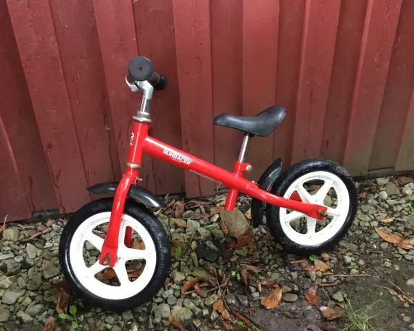 En röd stiga springcykel står lutad mot en röd trävägg. Den har vita fälgar och svarta däck.