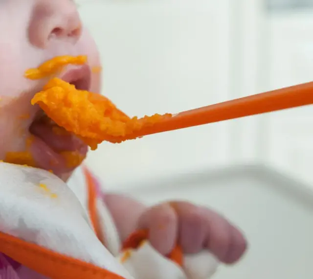 En bebis får mat på en sked, med orange puré runt munnen. En liten mage växer, och snart är det dags för mer mat.