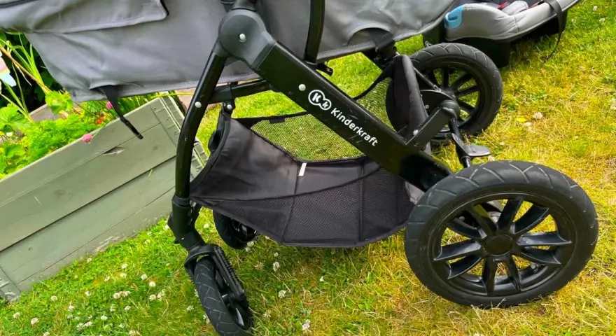 En barnvagn från Kinderkraft 3 in 1 står på gräset. Den har en svart ram, grå tygdelar och en bilbarnstol.
