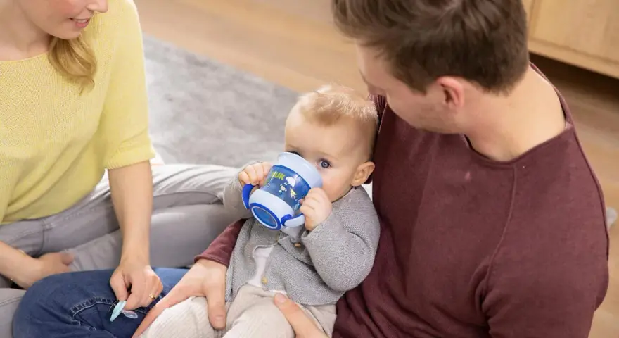 En bebis dricker ur en blå nuk magic cup medan föräldrarna tittar på.