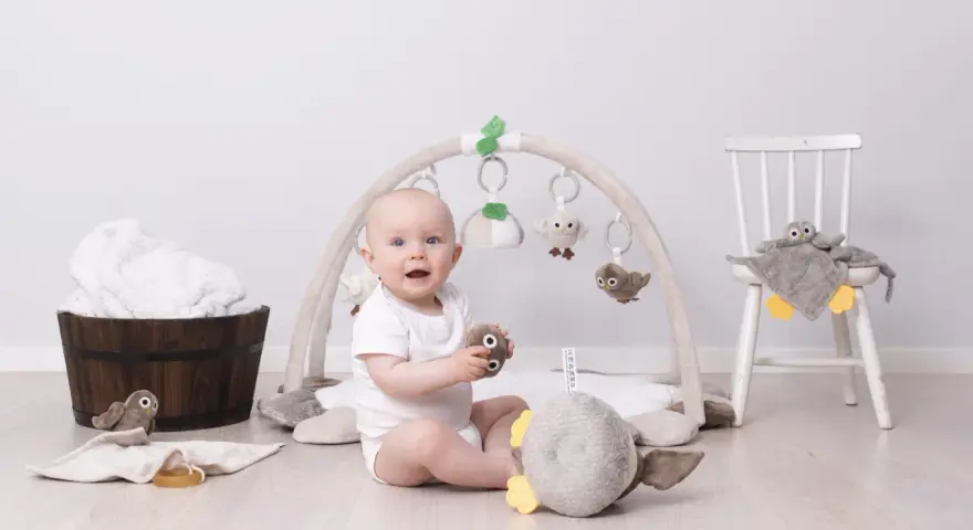Glad baby leker under en babygym båge med mjuka fågelleksaker. En stol med en uggla och en träbalja med handdukar i bakgrunden.