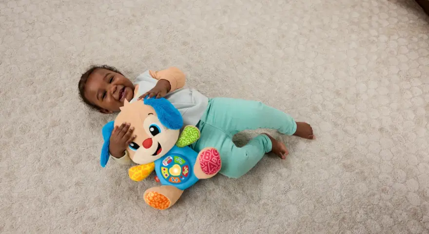 Ett litet barn skrattar och kramar sin Fisher Price hund på en ljus matta. Hunden har blå öron och en färgglad mage med knappar.