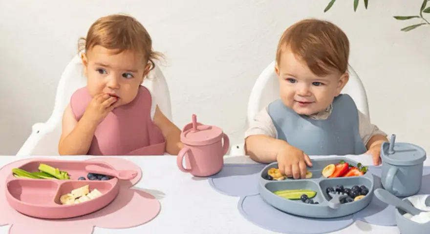 Två små barn äter från varsin tallrik ur ett barnservis i silikon. En rosa och en blå.