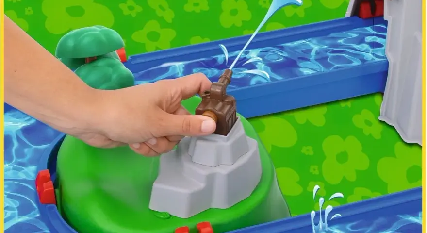 En hand aktiverar en vattenpump på en Aquaplay mountain lake-lekplats, som sprutar vatten genom en blå kanal.