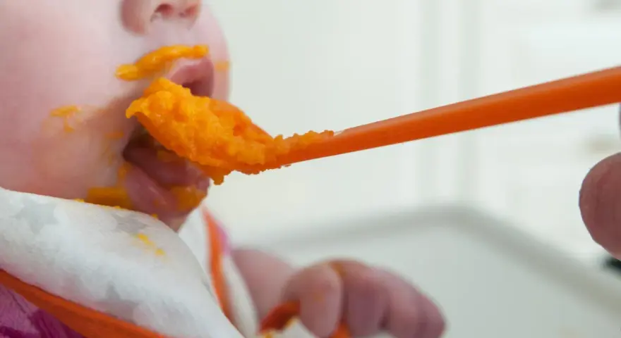 En bebis får mat på en sked, med orange puré runt munnen. En liten mage växer, och snart är det dags för mer mat.