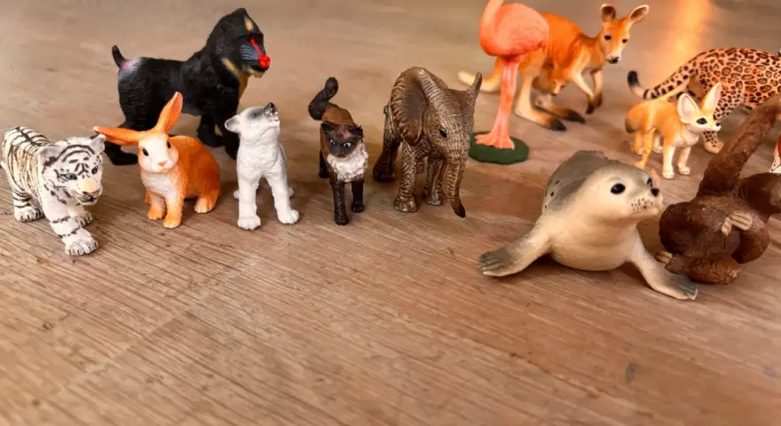En samling schleich vilda djur leksaker på ett trägolv: tiger, apa, kanin, hundvalp, katt, elefant, flamingo, känguru, fennek, säl och en apa.
