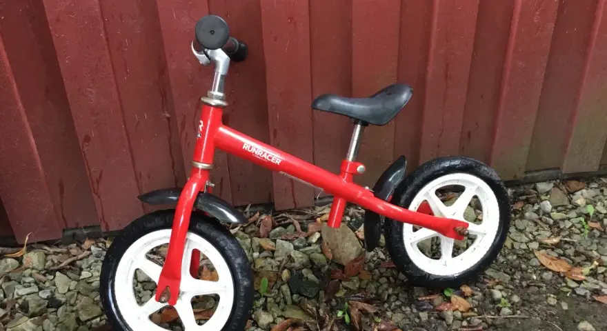 En röd stiga springcykel står lutad mot en röd trävägg. Den har vita fälgar och svarta däck.