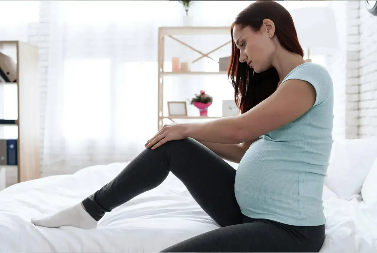 Gravid kvinna sitter p&aring; s&auml;ngen och gnuggar sitt ben, kanske p&aring; grund av myrkrypningar i benen gravid.