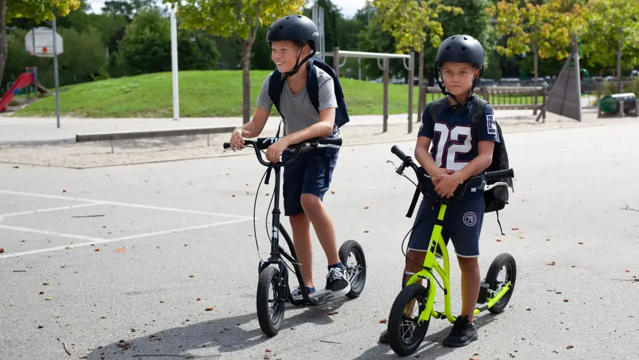 Två pojkar med hjälm och ryggsäckar kör stiga springcykel på skolgården.