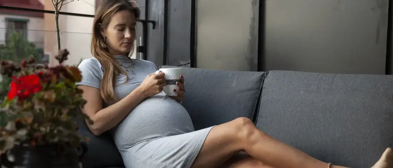 Gravid kvinna i tredje trimestern vilar på en soffa med en kopp i handen.