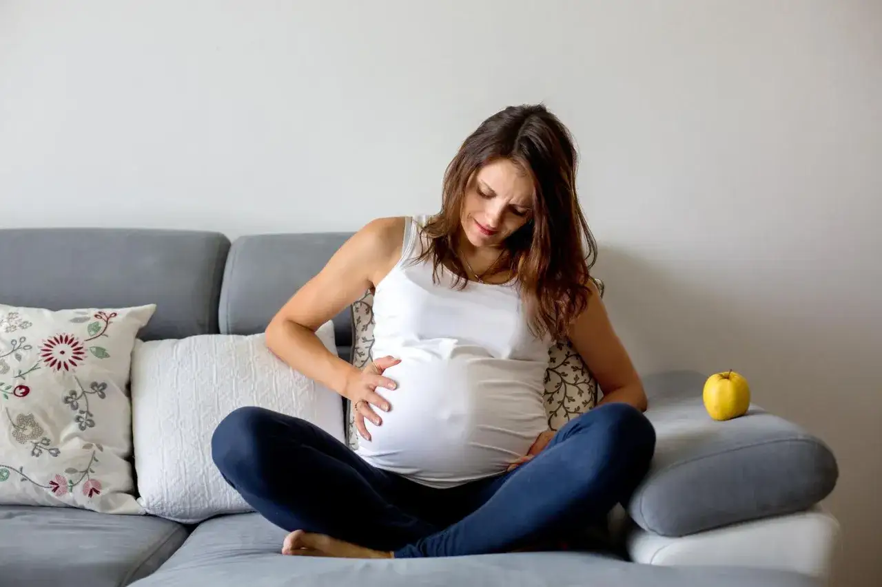 Gravid vecka 34, en kvinna sitter på en soffa och håller om sin mage. Familjeliv närmar sig.