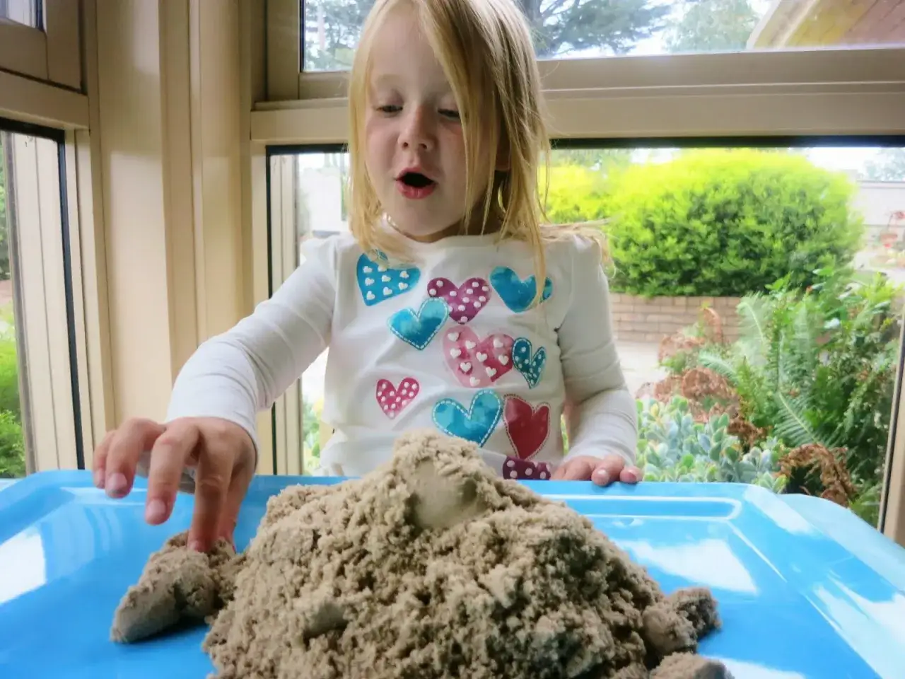 Liten flicka leker med kinetisk sand p&aring; ett bl&aring;tt fat. Hon bygger en sandh&ouml;g med sina fingrar.